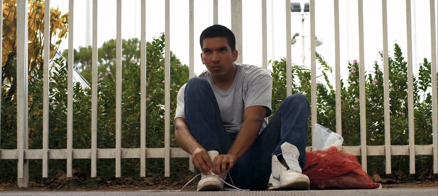 El Deportado production image: a young man sits beside a white metal fence, tying his shoelaces